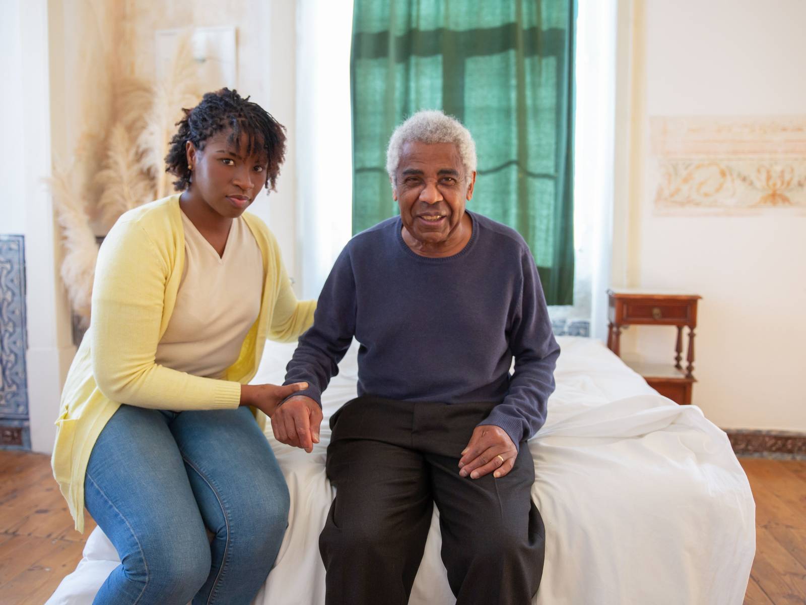 A woman sitting on a bed beside an older man, holding his hand