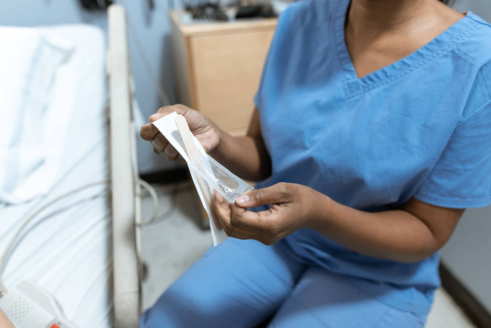 A healthcare worker in blue scrubs opening a packaged tongue depressor beside a hospital bed